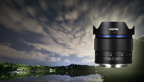 LAOWA ultra-wide angle lens with blue accent ring positioned against a nighttime landscape with reflective water and dramatic cloudy sky.