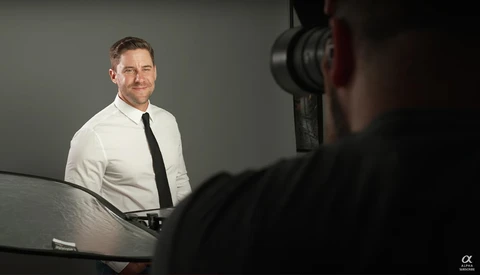 Man in white dress shirt and black tie seated at table, photographed from across the room during interview or portrait session.