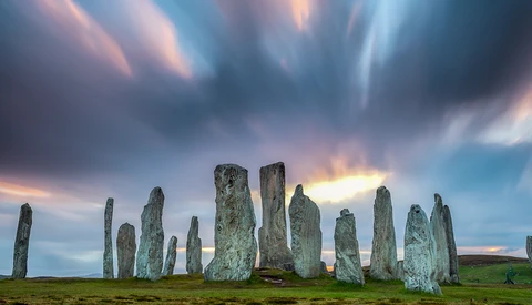 Ancient standing stones arranged in a circle on a grassy landscape beneath a dramatic twilight sky.