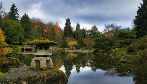 Stone lantern on moss-covered rock beside calm pond reflecting autumn foliage and evergreen trees.