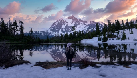 Hiker standing on snowy shore facing snow-capped mountain reflected in still lake at sunset.