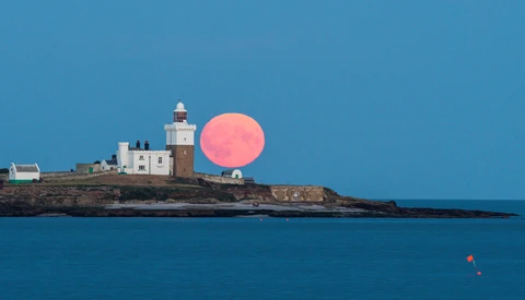 Lighthouse on rocky headland with oversized pink moon rising behind it against blue sky and sea.
