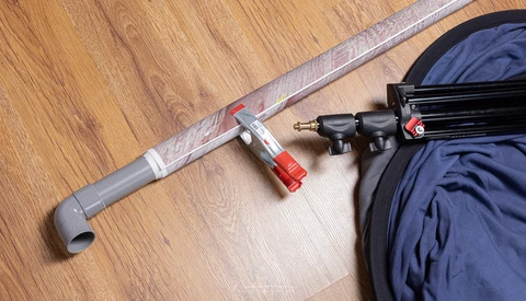 Tripod arm with red clamp and mounting hardware laid out on wooden floor next to camera bag.