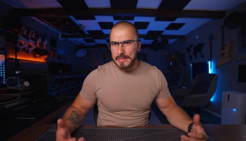 Man in beige t-shirt seated at desk in dimly lit room with blue and orange accent lighting.