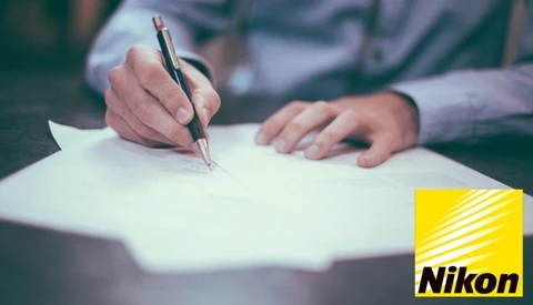 Person in light blue shirt writing on white paper with a pen at a desk.