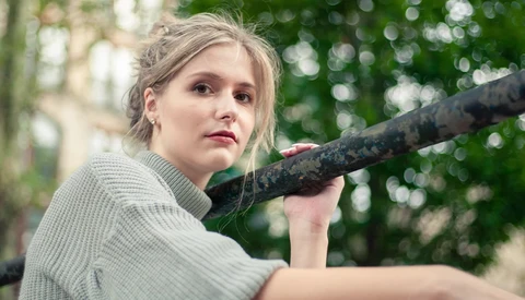 Young woman in light sweater holding weathered branch over shoulder, looking toward camera with serious expression.