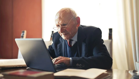 Older man wearing glasses and dark blazer working on laptop at desk in warm-lit interior space.