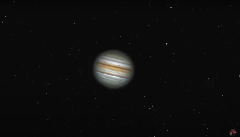Jupiter captured through a telescope showing distinctive cloud bands and atmospheric detail against a starfield.