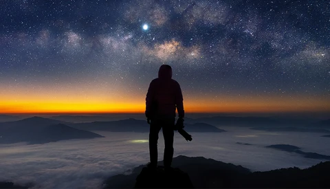 Silhouetted hiker standing on a mountain peak at sunrise, gazing toward a starry sky and Milky Way galaxy.