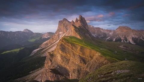 Dramatic alpine peak with steep rock faces and green meadows in a mountain valley below at golden hour.