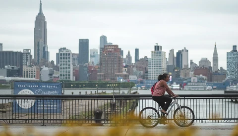 Cyclist riding a fixed-gear bike on a waterfront promenade with Manhattan skyline in background.