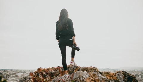 Woman standing on rocky outcrop holding camera, wearing dark clothing against overcast sky.