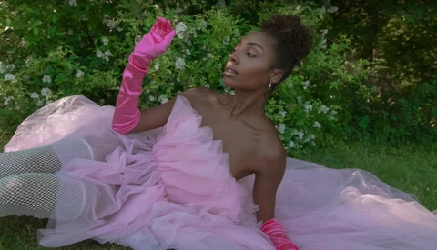 Woman in lavender tulle gown with pink arm warmers posed against green foliage backdrop.