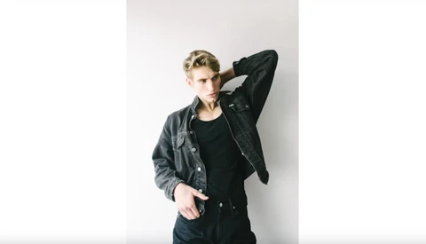 Male model in black denim jacket and shirt against white wall, hand raised to head.