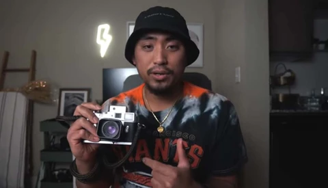 Photographer displaying a vintage rangefinder camera while wearing a black bucket hat and dark graphic shirt.