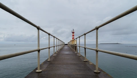 Long wooden pier extending into calm water toward a red and white striped lighthouse in the distance.