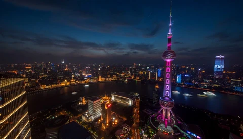 Illuminated Oriental Pearl Tower rises above Shanghai's skyline at dusk, surrounded by glowing city lights and a river.