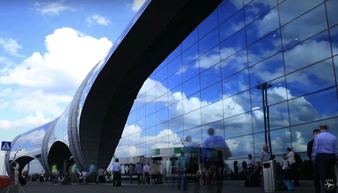 Modern curved glass building facade reflecting blue sky with people gathering at entrance below.