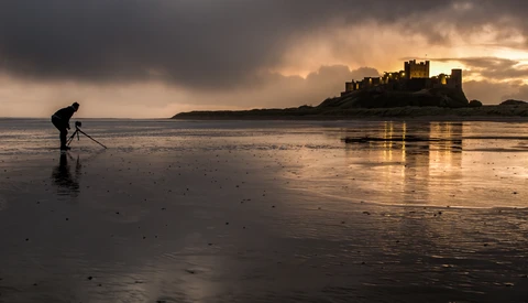 Photographer with tripod on wet beach at sunset, medieval castle ruins visible across the water.