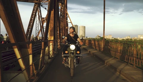 Motorcyclist riding across an industrial steel bridge toward a city skyline at golden hour.
