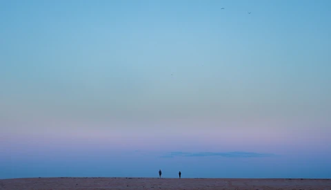 Two silhouetted figures standing on a vast, flat beach during twilight with a purple and blue sky.