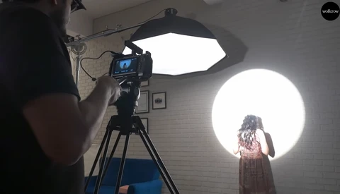 Filmmaker operating camera on tripod while subject stands before large circular backlit panel in studio.