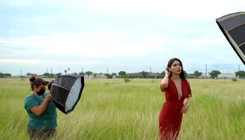 Photographer holding softbox reflector while model in red dress poses in open field.