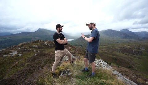 Two men standing on a rocky hilltop with mountains and valley landscape visible in the distance.