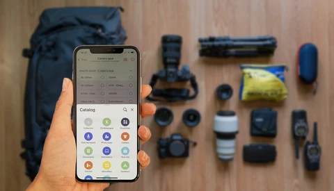 Photographer's hand holding smartphone displaying a catalog app interface with various photography equipment laid out on wooden surface.