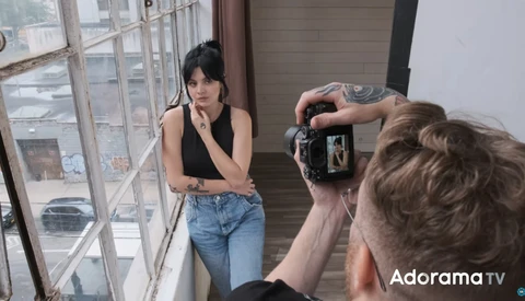 Photographer holding a camera on a tripod while shooting a portrait of a woman posing by a window.