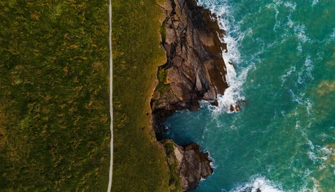 Aerial view of rocky coastal cliffs meeting turquoise ocean waves, with forested terrain and a straight road cutting through green landscape.