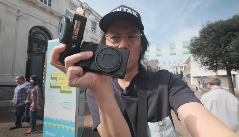 Photographer wearing a black cap holds a compact digital camera toward the camera in an urban plaza.