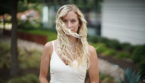 Woman with long blonde wavy hair wearing a white sleeveless top, photographed outdoors with soft natural light.
