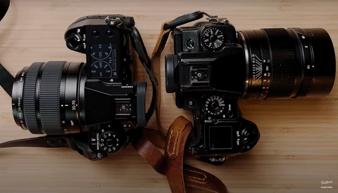 Two black film SLR cameras with attached lenses on a wooden surface, connected by a brown leather strap.