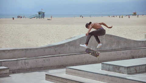 Shirtless skateboarder performing a trick on concrete ledges with beach and lifeguard tower in background.