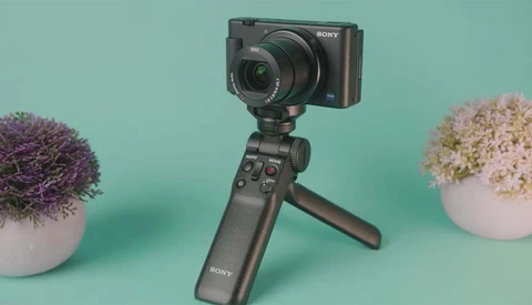 Sony compact camera mounted on Sony tripod grip with extended legs against turquoise background.
