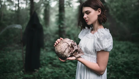 Woman in white period dress holding a mushroom in a dark forest clearing.
