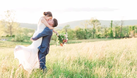 Groom lifting bride in a spin across an open meadow with wildflower bouquet.