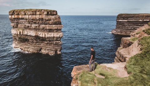 Person standing on grassy cliff edge between two large rock formations jutting from the sea.