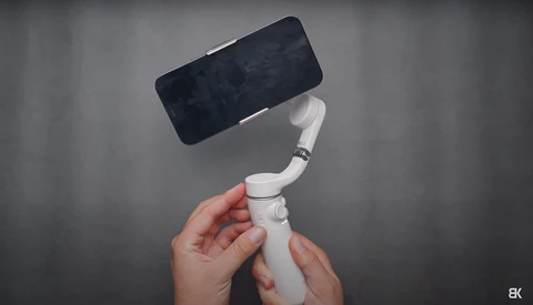 Hands holding a white articulating phone mount with a black smartphone attached against a gray background.
