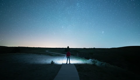 Silhouetted figure standing on a boardwalk, illuminated by headlamp light, facing a starry night sky over a dark landscape.