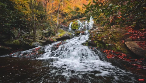 Cascading waterfall flowing through autumn forest with moss-covered rocks and fall foliage.