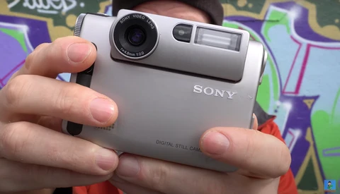 Person holding a silver Sony digital still camera with a compact design and retractable lens.