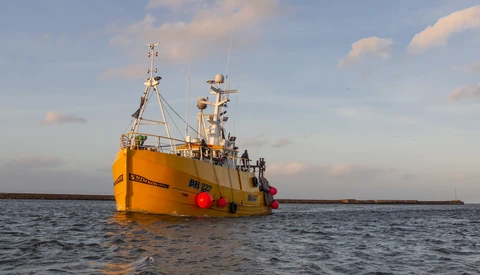 Yellow fishing vessel with red bumpers floating on calm blue water under partly cloudy sky.