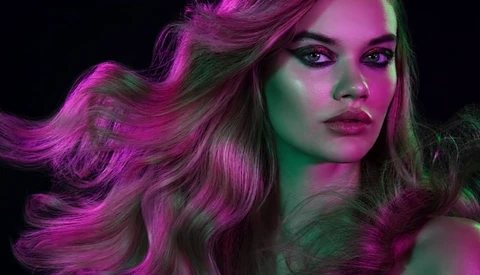Woman with long wavy hair illuminated by magenta and green gel lighting against a black background.