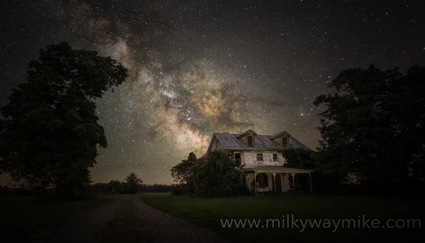 Abandoned two-story farmhouse silhouetted against the Milky Way galaxy stretching across a starry night sky.