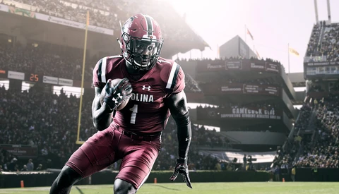 Football player in maroon uniform running with ball in stadium during game.
