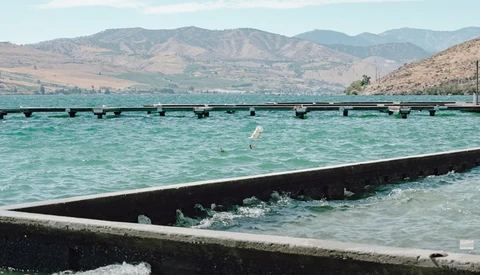 Turquoise water enclosed by black fish farm nets with arid mountains visible across the bay.