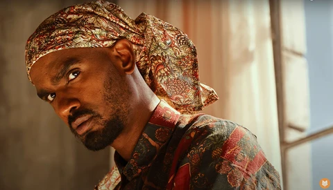 Man wearing patterned headwrap and red ornate shirt, photographed in warm indoor lighting.