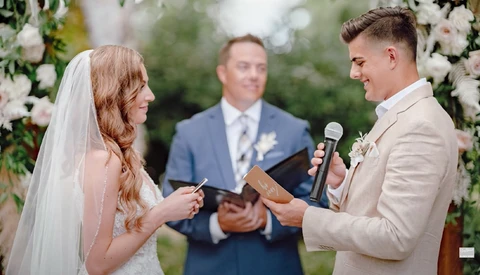 Groom and bride exchange vows during outdoor wedding ceremony with officiant in background.